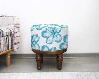 May include: A small, round wooden stool with a white top patterned with large, turquoise floral designs. The stool has four turned legs. The design is set against a white wall. The style is a blend of rustic and floral.