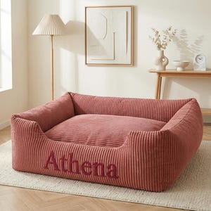 May include: A dusty rose corduroy dog bed with the name "Athena" embroidered in a darker shade. The bed has a rectangular shape with raised sides for comfort. The bed is placed on a light-colored rug.