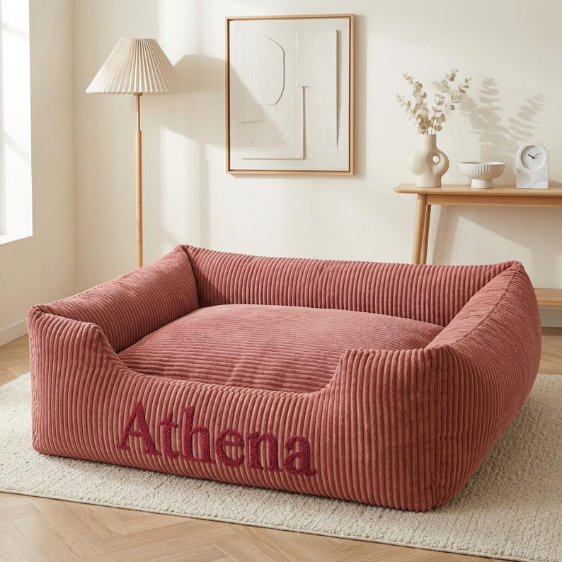 May include: A dusty rose corduroy dog bed with the name "Athena" embroidered in a darker shade. The bed has a rectangular shape with raised sides for comfort. The bed is placed on a light-colored rug.