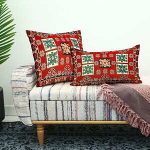 May include: Two decorative throw pillows with a red background and a geometric pattern in green, white, and gold. The pillows are on a striped bench with a pink blanket and a potted plant.