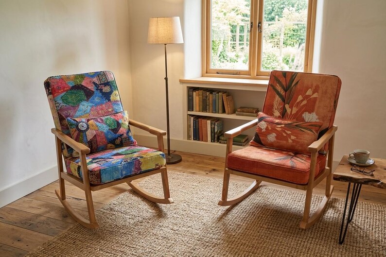 May include: Two wooden rocking chairs with patterned cushions. One chair features a patchwork design, the other an orange floral pattern. A floor lamp and a small side table are also visible. The chairs are in a room with a window.
