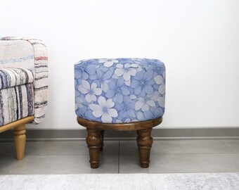 May include: A small, round wooden stool with a floral pattern. The cushioned top features a blue and white flower design. The stool has four turned wooden legs and is set against a white wall.