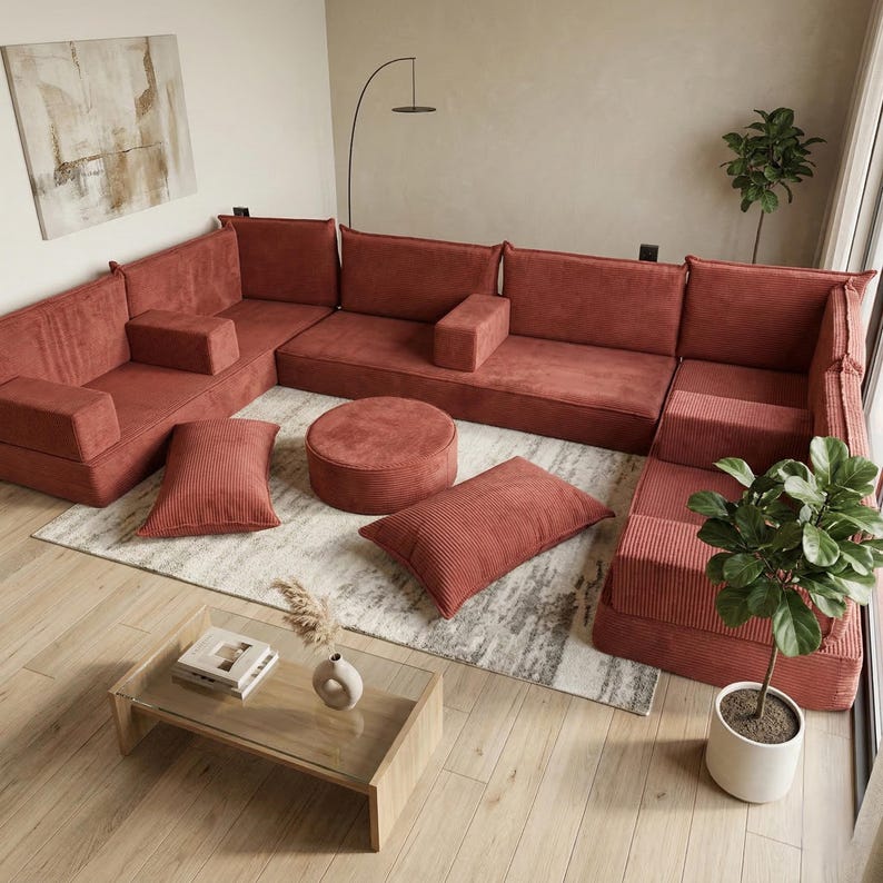 May include: A modular, rust-colored corduroy sectional sofa with matching throw pillows and a round ottoman. The sofa is arranged in an L-shape on a patterned rug, with a glass-top coffee table and potted plants nearby.