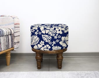 May include: A square ottoman with a dark blue floral pattern featuring white hibiscus flowers and tropical leaves. The ottoman sits on a dark wooden base with four turned legs. The design evokes a tropical theme.