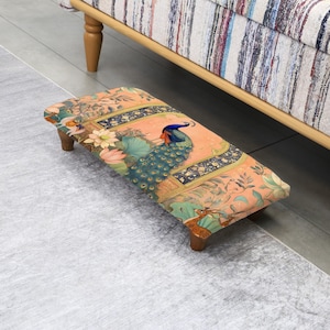 May include: A rectangular footstool with a colorful peacock design. The stool has a peach-colored background with a peacock in the center, surrounded by floral and leaf patterns. The legs are made of wood.