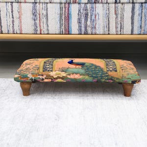 May include: A rectangular footstool with a peacock design. The stool has a peach-colored background with a peacock in the center, surrounded by floral and architectural elements. It rests on four wooden legs and is positioned under a sofa.