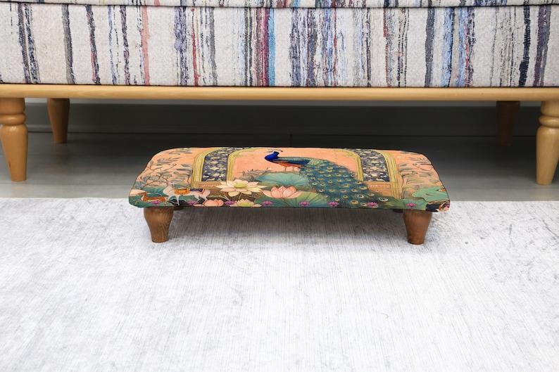 May include: A rectangular footstool with a peacock design. The stool has a peach-colored background with a peacock in the center, surrounded by floral and architectural elements. It rests on four wooden legs and is positioned under a sofa.