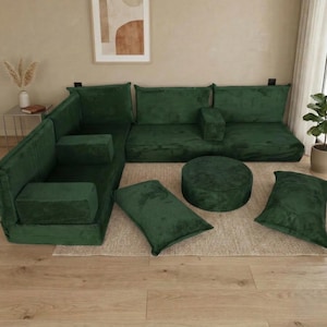 Op de afbeelding: Een modulaire hoekbank van bosgroen corduroy met bijpassende kussens en een ronde poef. De bank is in een L-vorm gerangschikt, met extra kussens en een ronde poef. De meubels staan op een neutraal gekleurd tapijt.