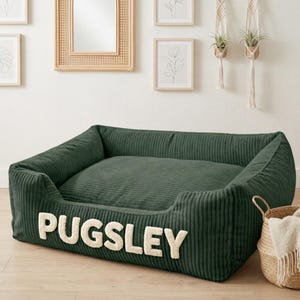 May include: A dark green corduroy dog bed with the name "PUGSLEY" in cream-colored letters. The bed has a rectangular shape with raised sides and a soft, cushioned interior. A woven basket with a white blanket is next to the bed.