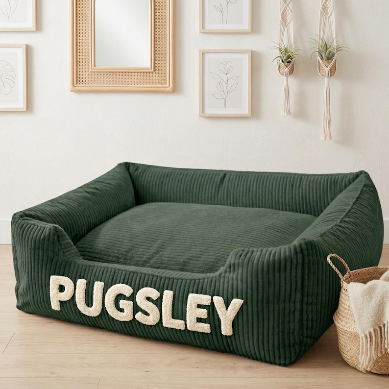 May include: A dark green corduroy dog bed with the name "PUGSLEY" in cream-colored letters. The bed has a rectangular shape with raised sides and a soft, cushioned interior. A woven basket with a white blanket is next to the bed.