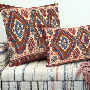 Pode incluir: Duas almofadas decorativas com fundo creme e um padrão geométrico em tons de azul, vermelho e castanho. As almofadas são colocadas num sofá estampado, criando uma atmosfera acolhedora e convidativa. As almofadas adicionam um toque de cor e padrão à divisão.