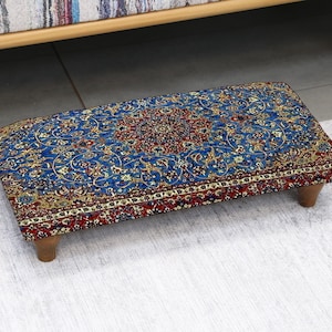 Peut inclure: Un pouf rectangulaire avec un motif de tapis détaillé en bleu, rouge et or. Le pouf a de courts pieds en bois. Le tapis présente un médaillon central et des motifs floraux.