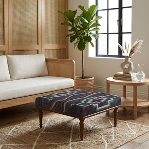 Puede incluir: Un otomán cuadrado con tapicería de tela azul oscuro y un patrón geométrico en blanco, gris y rojo. El otomán se apoya sobre cuatro patas de madera torneada con un acabado marrón medio, proporcionando una base estable para el cojín.