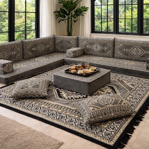 Op de afbeelding: Een grijze hoekbank met kussens met patronen en een bijpassende poef. De bank staat op een groot vloerkleed met een zwarte franje. Decoratieve kussens en een dienblad met eten maken de woonkamer compleet.