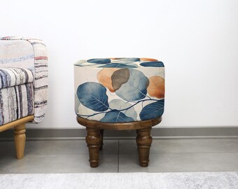 May include: A small, round wooden stool with a cushioned top. The cushion has a watercolour design of blue, orange, and grey leaves on a cream background. The stool has four turned legs and is in front of a white wall.
