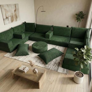 May include: A modular green corduroy sectional sofa with matching pillows and a round ottoman. The sofa is arranged in an L-shape on a light-colored rug. A glass-top coffee table and potted plant are in the foreground.