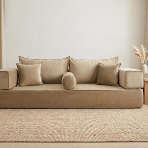Könnte beinhalten: Ein beiges Modulsofa mit mehreren Kissen und einer zylindrischen Rolle. Das Sofa steht auf einem gewebten Jute-Teppich. Der Raum hat neutrale Töne, mit einem kleinen Beistelltisch aus Holz und einem Topf-Olivenbaum.