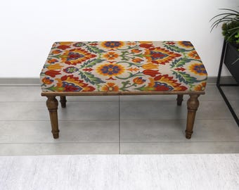 Puede incluir: Un otomán rectangular con un colorido estampado floral en rojo, naranja, amarillo y azul sobre un fondo crema. Las patas de madera son de color marrón oscuro. El otomán es un mueble decorativo.