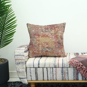 May include: A square cushion featuring a vintage rug pattern in red, beige, and brown tones. The cushion sits on a striped bench with wooden legs. A fringed throw blanket is also visible.