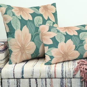 May include: Two decorative cushions with a floral design. The cushions display large peach-coloured flowers and green leaves on a teal background. They are placed on a patterned sofa.