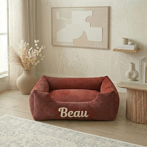 May include: A corduroy dog bed in a reddish-brown hue, with the name "Beau" embroidered in white. The bed has a rectangular shape with raised sides and a plush interior, designed for pet comfort.