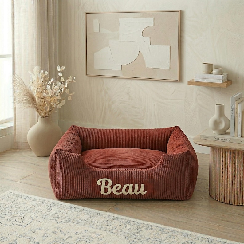 May include: A corduroy dog bed in a reddish-brown hue, with the name "Beau" embroidered in white. The bed has a rectangular shape with raised sides and a plush interior, designed for pet comfort.