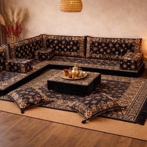 Puede incluir: Un sofá esquinero negro y dorado con estampado, con cojines a juego y una mesa de centro cuadrada. El sofá se encuentra sobre una alfombra estampada, con una alfombra de fibra natural debajo. Un juego de té decorativo está sobre la mesa de centro.