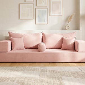 May include: A large, low-profile pink sofa with multiple cushions and a cylindrical bolster. The sofa has a minimalist design with square armrests. The room features framed artwork, a lamp, and a neutral-toned rug on a hardwood floor.