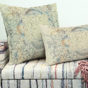 May include: Two decorative cushions with ornate, vintage-style patterns in beige, blue, and orange tones rest on a patterned sofa. The cushions feature a central medallion design and scrollwork.