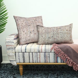 May include: Two decorative pillows with a vintage-style pattern rest on a striped bench. A matching throw with fringe and a potted palm tree complete the scene. The pillows and throw feature muted colors.
