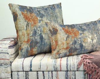 Può includere: Due cuscini decorativi con un design astratto nei toni del blu, arancione e grigio. I cuscini poggiano su un divano imbottito a righe. È visibile una coperta con frange.