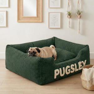 May include: A dark green corduroy dog bed with the name "PUGSLEY" embroidered in cream-colored letters. A tan pug dog is resting inside the bed. The bed is square-shaped with raised sides.