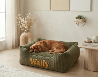 May include: A sleeping Cavalier King Charles Spaniel rests on a green corduroy dog bed. The bed has a rectangular shape with raised sides and the name "Wally" embroidered in gold thread. The bed is placed on a light-colored rug.