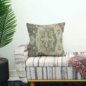 May include: A cushion featuring a muted, vintage-style rug pattern in grey and beige tones. It sits on a striped bench with a wooden frame. A potted palm tree is visible on the left.