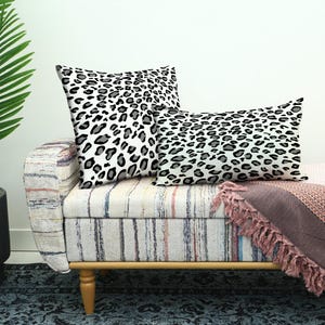 May include: Two decorative throw pillows with a black and gray leopard print pattern rest on a striped bench. A pink blanket with fringe is draped over the bench. A potted plant is in the background.