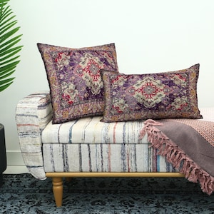 May include: Two decorative throw pillows with a Persian rug design in shades of purple, red, and gold rest on a striped bench. A matching throw blanket with fringe adds a touch of elegance. A potted palm tree is in the background.