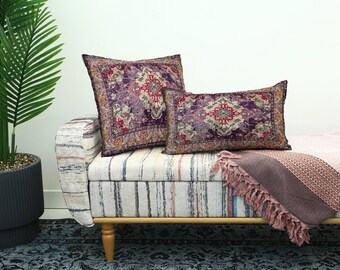 Op de afbeelding: Twee decoratieve sierkussens met een Perzisch tapijtontwerp in paars, rood en goud liggen op een gestreepte bank. Een bijpassende plaid met franjes voegt een vleugje elegantie toe. Op de achtergrond staat een palmboom in pot.
