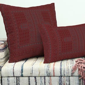 May include: Two burgundy cushions with a geometric design are placed on a striped sofa. The cushions are rectangular and square. The sofa has a multi-coloured striped pattern. The cushions are in a deep red hue.