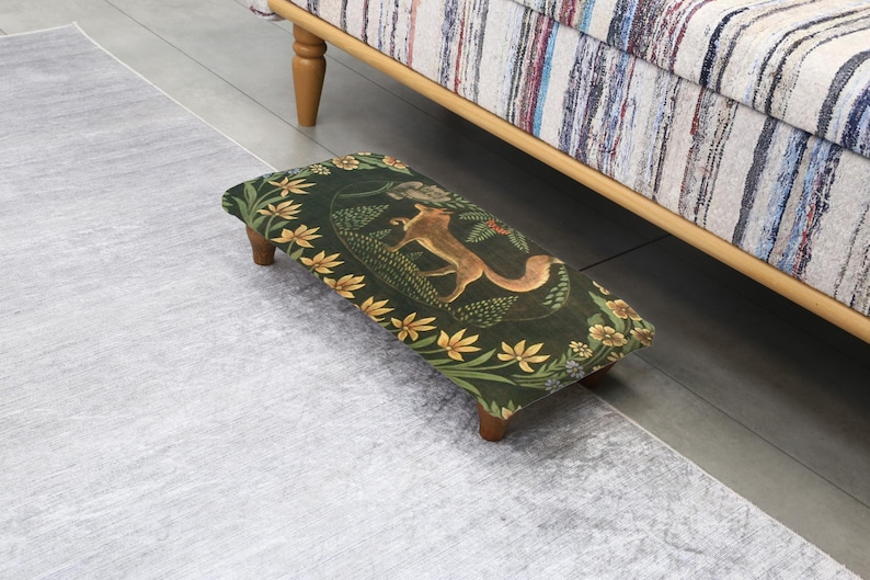 Op de afbeelding: Een rechthoekige voetenbank met een donkergroene stof met een vos en bloemenmotief. De voetenbank heeft houten poten en staat op een lichtgrijs tapijt. Het ontwerp bevat gele en witte bloemen.