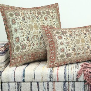 Puede incluir: Dos cojines decorativos con fondo crema y un detallado estampado floral en tonos rojos, marrones y azules. Los cojines están colocados sobre un sofá a rayas. También se ve una manta con flecos.