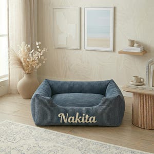 May include: A blue corduroy pet bed with the name "Nakita" embroidered in white. The bed has a rectangular shape with raised sides and a soft, cushioned interior. The bed is in a living room setting.