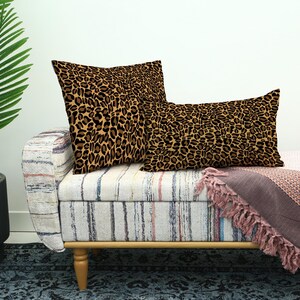 May include: Two decorative throw pillows with a brown and black leopard print pattern rest on a striped bench. A pink blanket with fringe is draped over the bench. A potted plant is in the background.