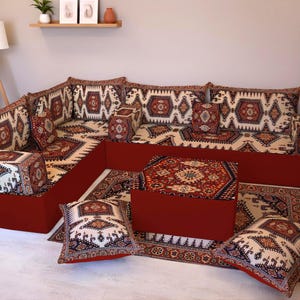 Puede incluir: Un sofá esquinero con base roja y cojines y almohadas estampados. Los cojines y almohadas presentan un fondo crema con un diseño geométrico repetitivo en tonos rojos, marrones y negros. Una alfombra y una otomana a juego completan el conjunto.