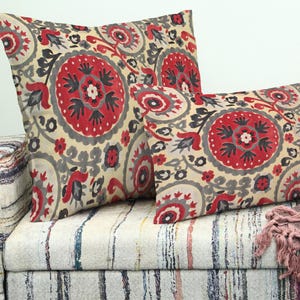 May include: Two decorative cushions with a red, grey, and cream floral pattern rest on a patterned sofa. The cushions feature a large, circular design with intricate details. The sofa has a striped fabric.