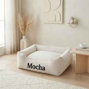 May include: A cream-colored dog bed with the word "Mocha" in black script. The bed has a corduroy texture and a rectangular shape with raised sides. The bed is placed on a light wood floor.