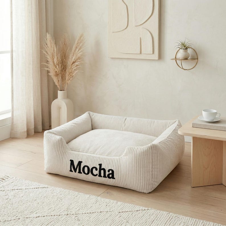 May include: A cream-colored dog bed with the word "Mocha" in black script. The bed has a corduroy texture and a rectangular shape with raised sides. The bed is placed on a light wood floor.