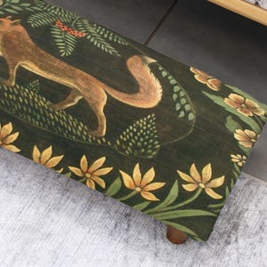 Op de afbeelding: Houten bank met een donkergroen oppervlak met een vosillustratie. De vos is bruin en bruin, omringd door groene bladeren en gele bloemen. De bank heeft een bloemenrand en houten poten.