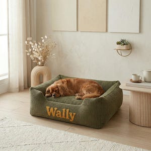 May include: A sleeping Cavalier King Charles Spaniel rests on a green corduroy dog bed. The bed has a rectangular shape with raised sides and the name "Wally" embroidered in gold thread. The bed is placed on a light-colored rug.