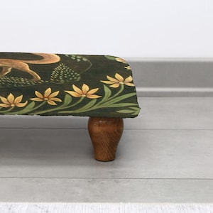 Op de afbeelding: Een decoratieve voetenbank met een donkergroene stoffen bovenkant met een vos en bloemenmotief. De randen zijn versierd met gele bloemen en groene bladeren. De kruk heeft een houten basis en poten.