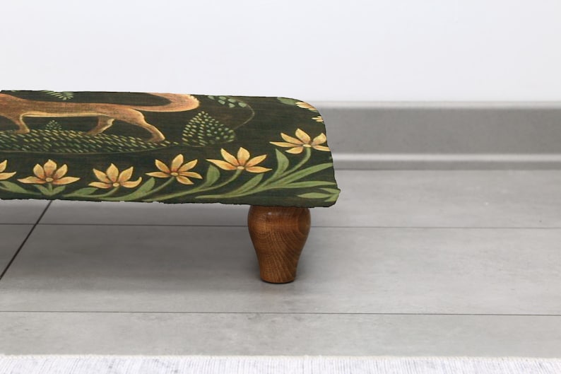 Op de afbeelding: Een decoratieve voetenbank met een donkergroene stoffen bovenkant met een vos en bloemenmotief. De randen zijn versierd met gele bloemen en groene bladeren. De kruk heeft een houten basis en poten.